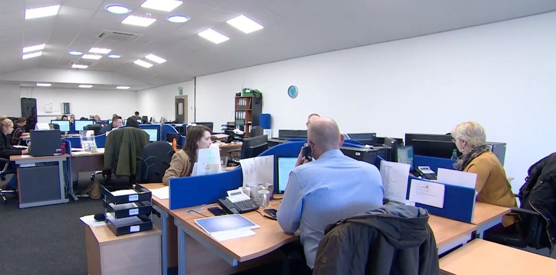 An open-plan office with several people working at desks with computers, phones, and paperwork. The space is well-lit with overhead lights, and there are blue desk dividers and office supplies visible.