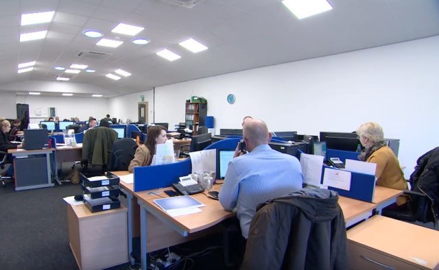 An open-plan office with several people working at desks with computers, phones, and paperwork. The space is well-lit with overhead lights, and there are blue desk dividers and office supplies visible.
