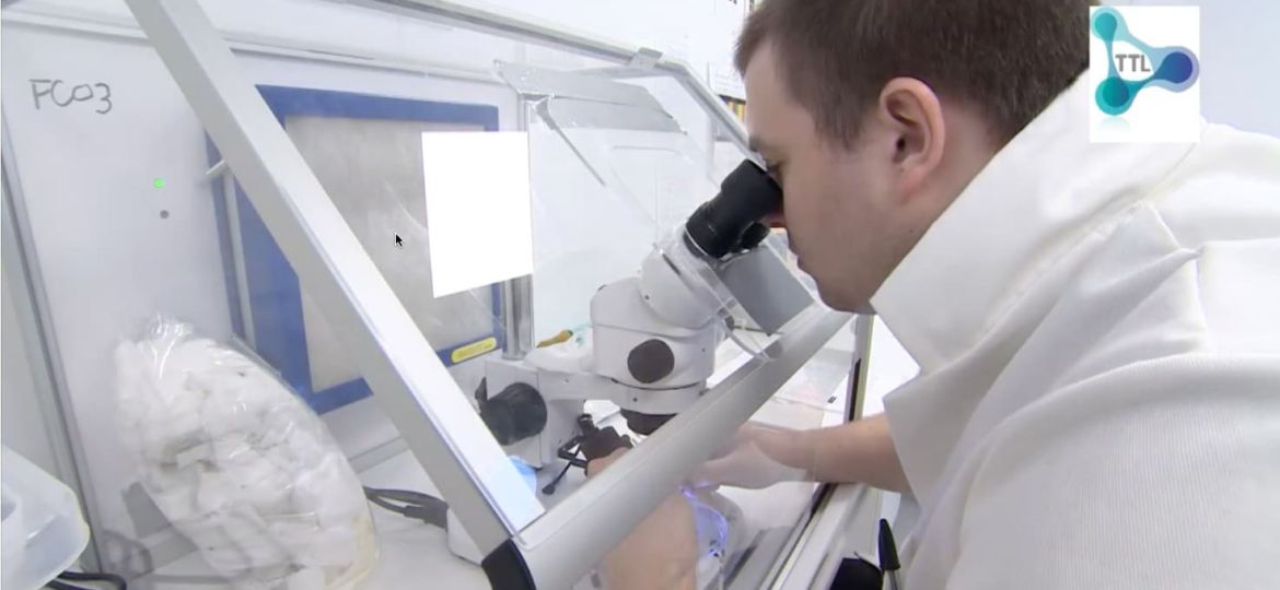 A person in a lab coat looks through a microscope inside a laboratory workstation, surrounded by scientific equipment and supplies, with a TTL logo visible in the top right corner.