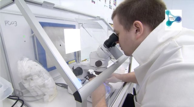 A person in a lab coat looks through a microscope inside a laboratory workstation, surrounded by scientific equipment and supplies, with a TTL logo visible in the top right corner.
