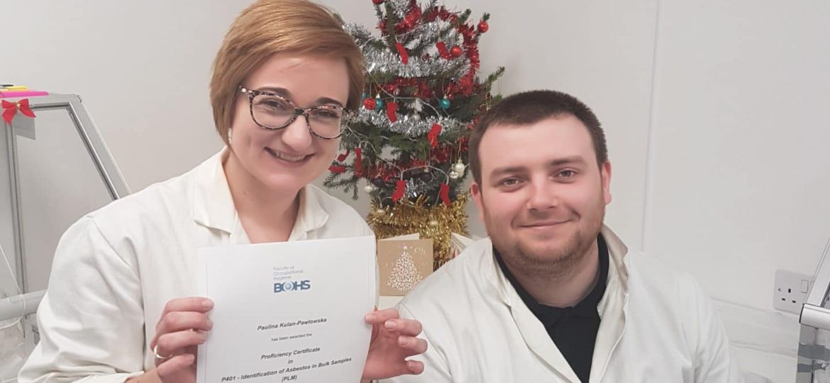 Two people in white lab coats pose in a laboratory. One person is holding a certificate and smiling at the camera. There is a small decorated Christmas tree in the background.