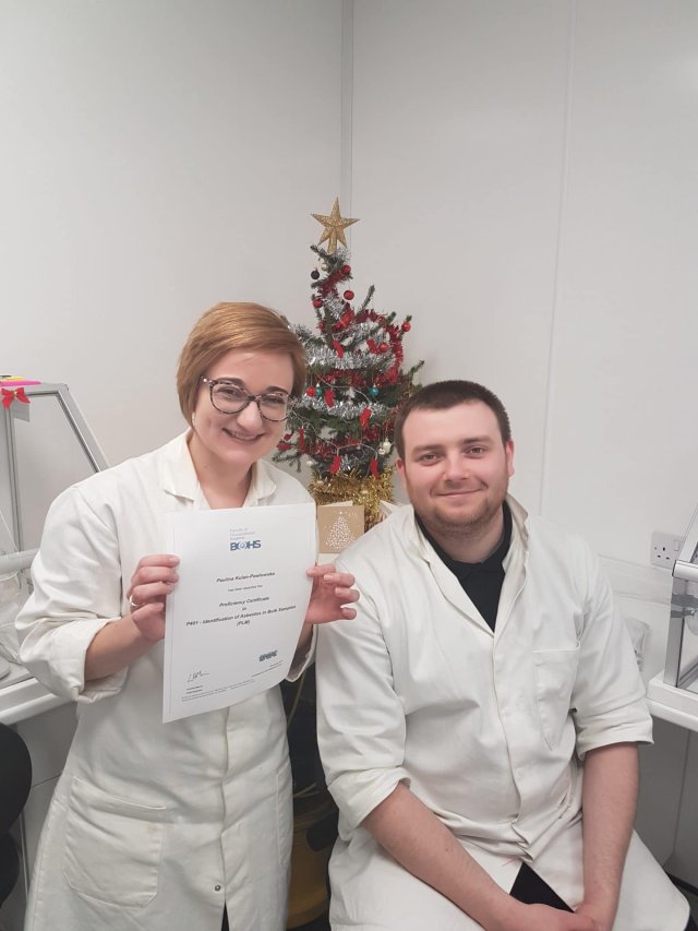 Two people in white lab coats pose in a laboratory. One person is holding a certificate and smiling at the camera. There is a small decorated Christmas tree in the background.