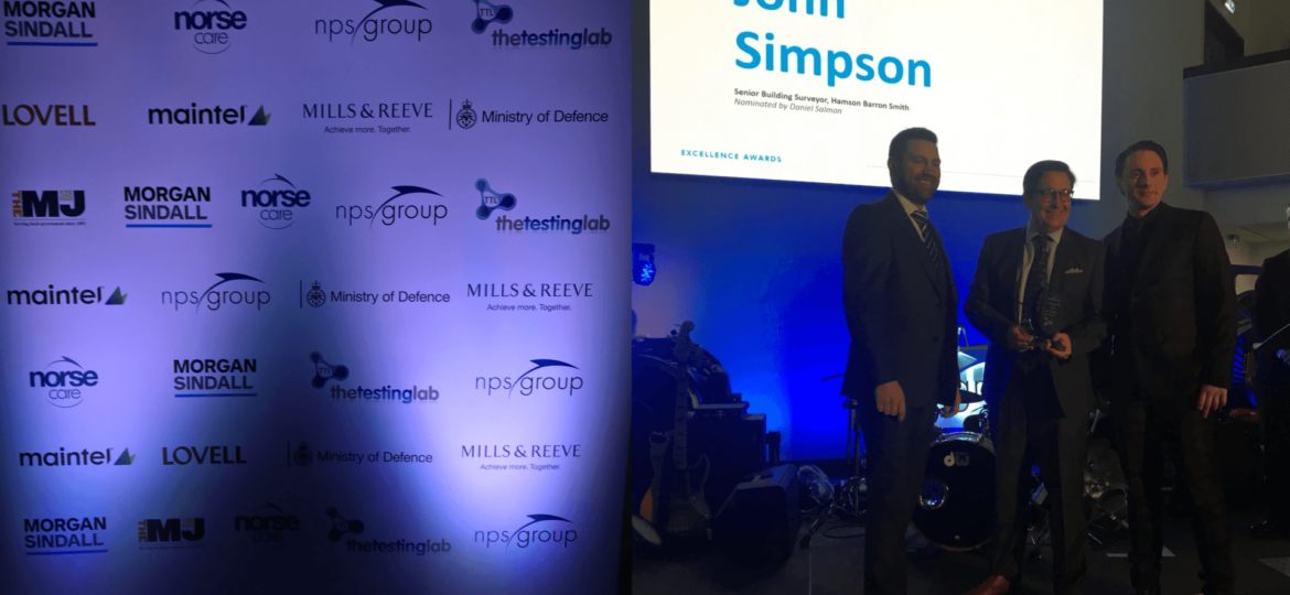 Three men stand on stage during an awards ceremony. Behind them, a large screen displays “John Simpson” next to smaller text, while a backdrop on the left shows logos of various sponsors including the Ministry of Defence.