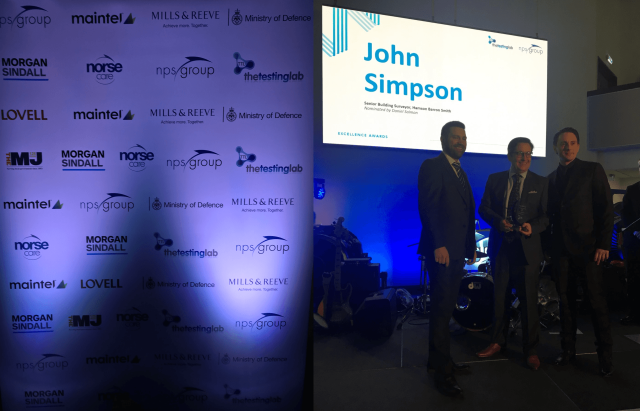 Three men stand on stage during an awards ceremony. Behind them, a large screen displays “John Simpson” next to smaller text, while a backdrop on the left shows logos of various sponsors including the Ministry of Defence.