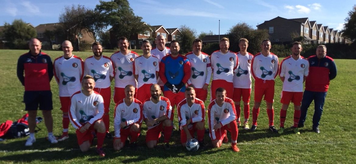 A football team in matching red and white kits poses for a group photo on a grassy pitch, with two coaches standing at each end. Houses and trees are visible in the background on a sunny day.