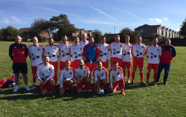 A football team in matching red and white kits poses for a group photo on a grassy pitch, with two coaches standing at each end. Houses and trees are visible in the background on a sunny day.