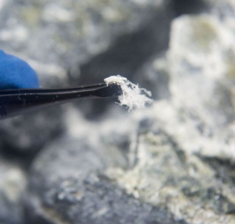 A close-up of tweezers holding a small, fibrous piece of asbestos, with grey rocks in the background. The tweezers are held by a hand wearing a blue glove.