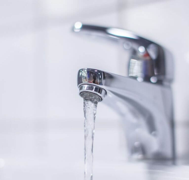 A close-up of a shiny chrome tap with water flowing from the spout, set against a blurred white tiled background.