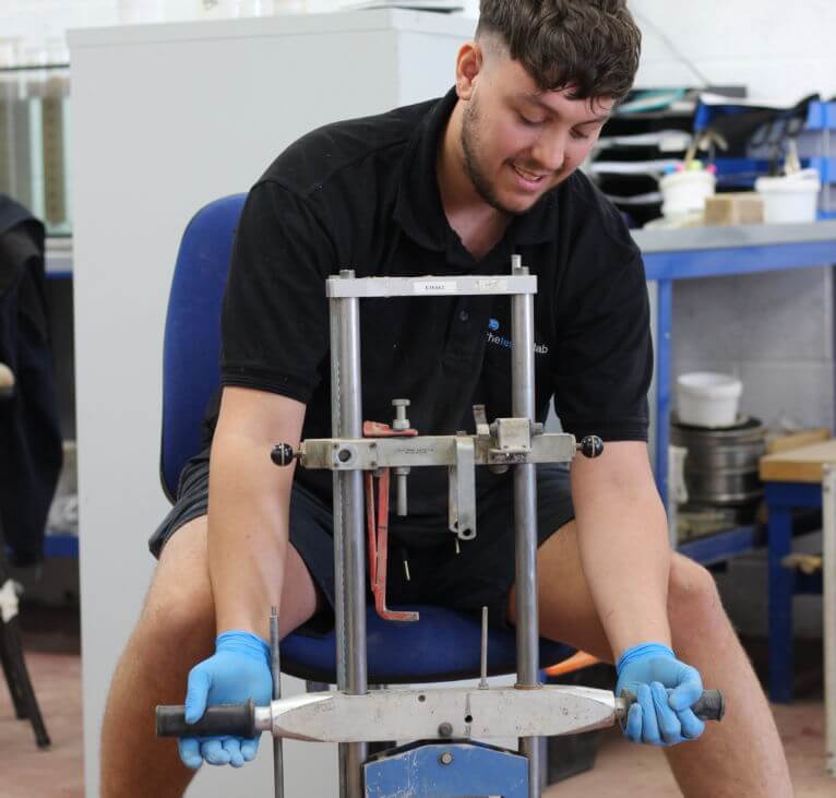 A man wearing blue gloves and a black shirt operates a manual laboratory press in a workshop, sitting on a chair and handling the equipment with both hands. Laboratory tools and shelves are visible in the background.