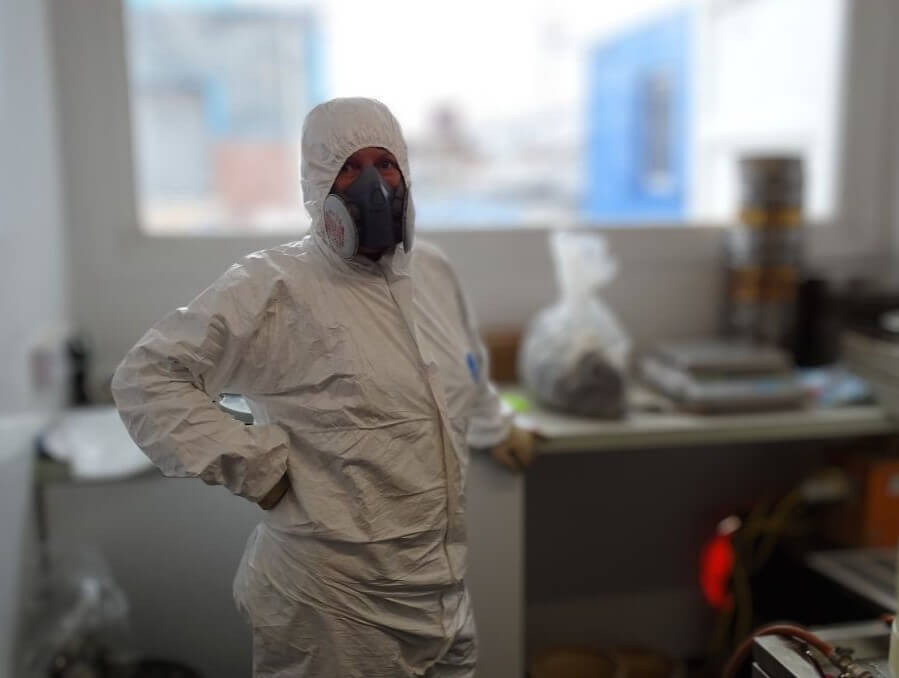 A person wearing a white hazmat suit, gloves, and a respirator stands in a laboratory, with equipment and supplies visible on benches in the background. A person wearing a white hazmat suit, gloves, and a respirator stands in a laboratory, with equipment and supplies visible on benches in the background.