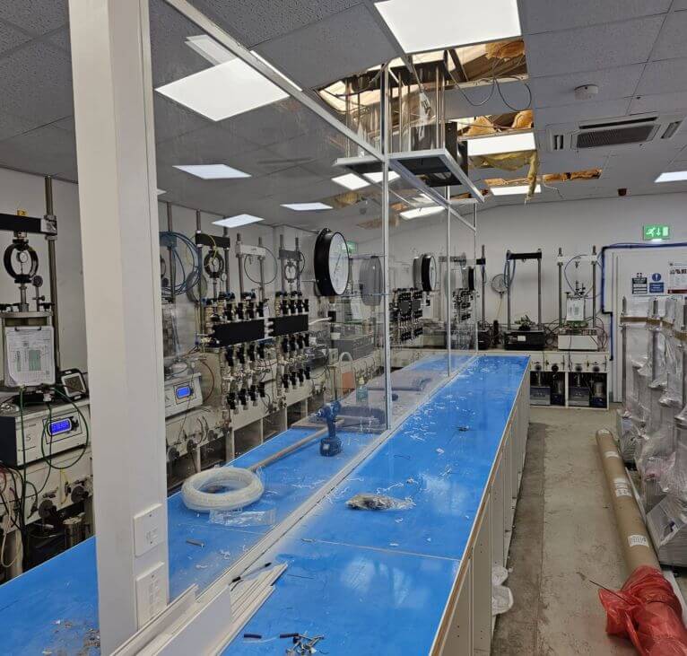 A laboratory with blue workbenches, scientific equipment, gauges on the walls, and a partially damaged ceiling exposing insulation. The room is well-lit with overhead lights and has various supplies and boxes along the right side.