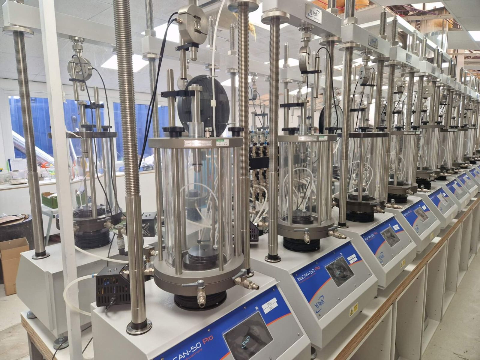 A row of automated laboratory testing machines with transparent chambers and digital displays, set up on a bench in a well-lit lab environment. The machines feature various metal components and control panels.