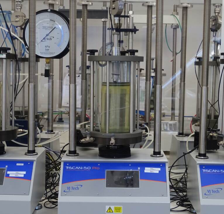 A laboratory triaxial testing machine with a transparent cylindrical cell, pressure gauges, and digital controls, used for soil mechanics experiments; wires and equipment are visible around it.