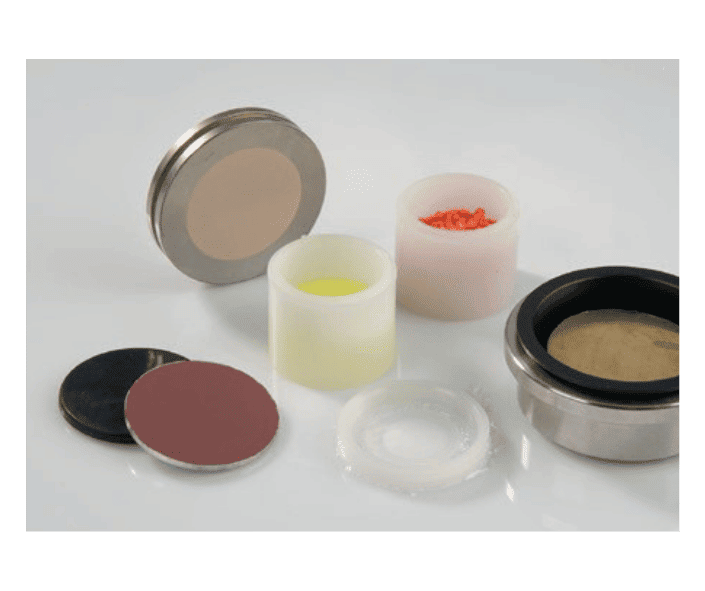 A variety of X-ray fluorescence sample cups, discs, and pellets are displayed on a white surface, including small containers with coloured powders and circular holders with different membrane materials.