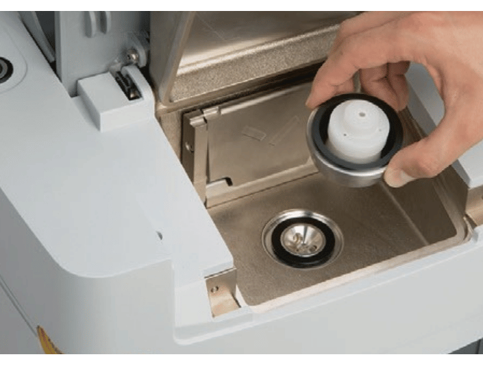 A hand removes a round water tank filter from a compartment in a coffee machine, revealing the inside of the water reservoir.