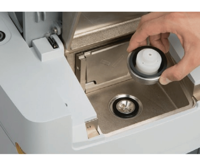 A hand removes a round water tank filter from a compartment in a coffee machine, revealing the inside of the water reservoir.